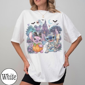 May include: A white short-sleeve t-shirt featuring a graphic print of two blue and pink alien characters in Halloween costumes. One character is dressed as a pumpkin, the other as a mummy. The background shows a castle, bats, and balloons.