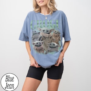 Puede incluir: Una camiseta azul claro con la palabra "GROGU" en letras verdes sobre un gr&aacute;fico de Grogu de The Mandalorian. El gr&aacute;fico muestra a Grogu en varias poses. La camiseta la lleva una persona que lleva pantalones cortos negros.