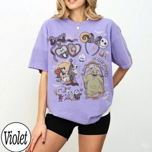 May include: A violet-colored t-shirt featuring a graphic design with characters from "The Nightmare Before Christmas." The design includes Jack Skellington, Sally, and Oogie Boogie, along with other Halloween-themed elements. The shirt has short sleeves.