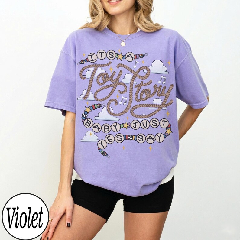 May include: A violet short-sleeve t-shirt featuring a graphic print with the text "ITS A Toy Story BABY JUST YES SAY". The design includes rope-style lettering, colorful beads, stars, musical notes, and white cloud shapes on the front.