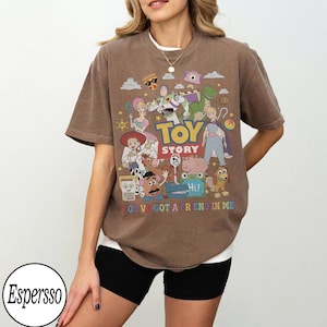 May include: A brown short-sleeve t-shirt featuring a colorful graphic print of Toy Story characters including Buzz Lightyear, Woody, and Jessie. The text reads TOY STORY and YOU'VE GOT A FRIEND IN ME, accompanied by various small icons like a pizza slice.