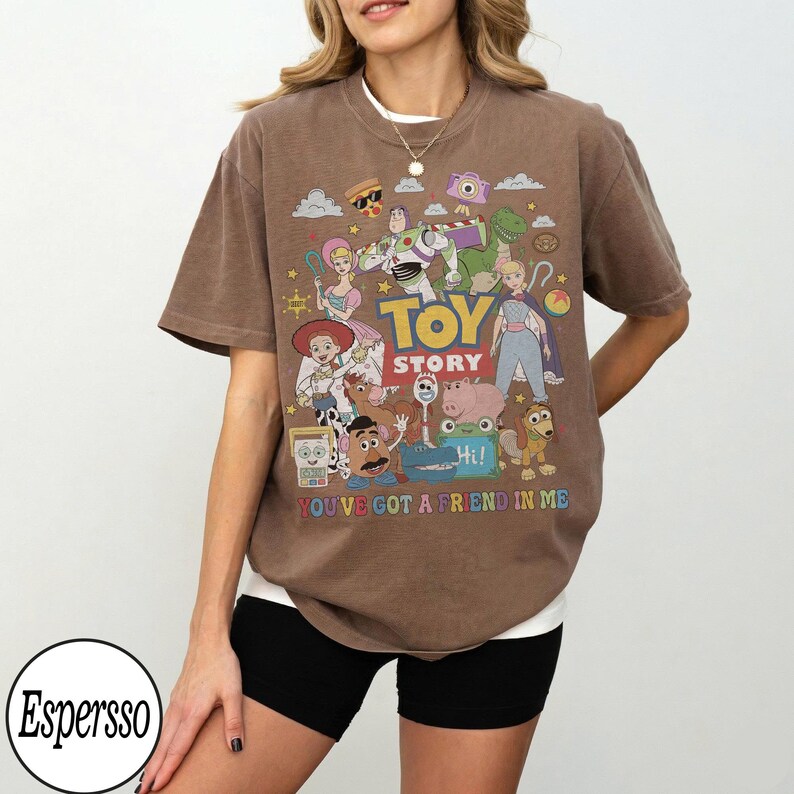 May include: A brown short-sleeve t-shirt featuring a colorful graphic print of Toy Story characters including Buzz Lightyear, Woody, and Jessie. The text reads TOY STORY and YOU'VE GOT A FRIEND IN ME, accompanied by various small icons like a pizza slice.
