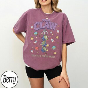 Puede incluir: Una camiseta de manga corta color baya con un estampado gr&aacute;fico que incluye el texto THE CLAW y THE MYSTIC PORTAL AWAITS. El dise&ntilde;o muestra tres alien&iacute;genas verdes, cohetes, planetas y estrellas en una ilustraci&oacute;n espacial en el frente.
