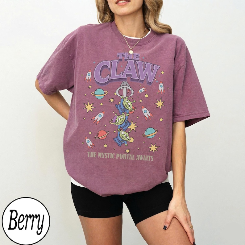 Puede incluir: Una camiseta de manga corta color baya con un estampado gr&aacute;fico que incluye el texto THE CLAW y THE MYSTIC PORTAL AWAITS. El dise&ntilde;o muestra tres alien&iacute;genas verdes, cohetes, planetas y estrellas en una ilustraci&oacute;n espacial en el frente.