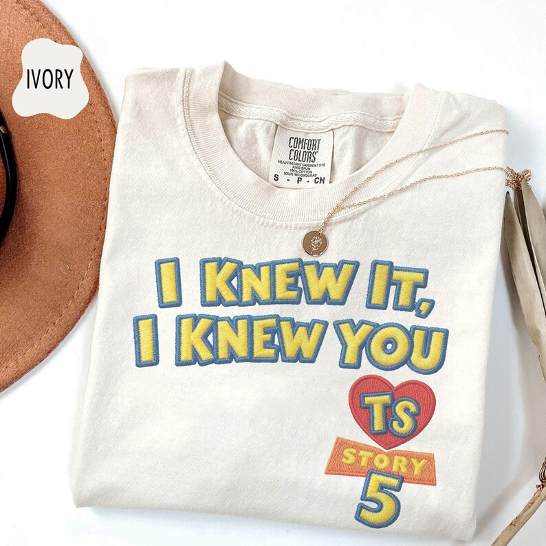 Puede incluir: Una camiseta de color marfil con el texto bordado I KNEW IT, I KNEW YOU en amarillo con contornos azules. Debajo del texto hay un coraz&oacute;n rojo que contiene las letras TS, con la palabra STORY y el n&uacute;mero 5 en amarillo debajo del coraz&oacute;n.