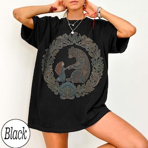 以下が含まれることがあります： 黒いTシャツ。円形のデザインで、クマと女の子が手をつないでおり、花のモチーフとアザミで囲まれています。デザインは茶色、青、緑の色合いを使用しています。「Black」の文字が下部の白い円の中にあります。