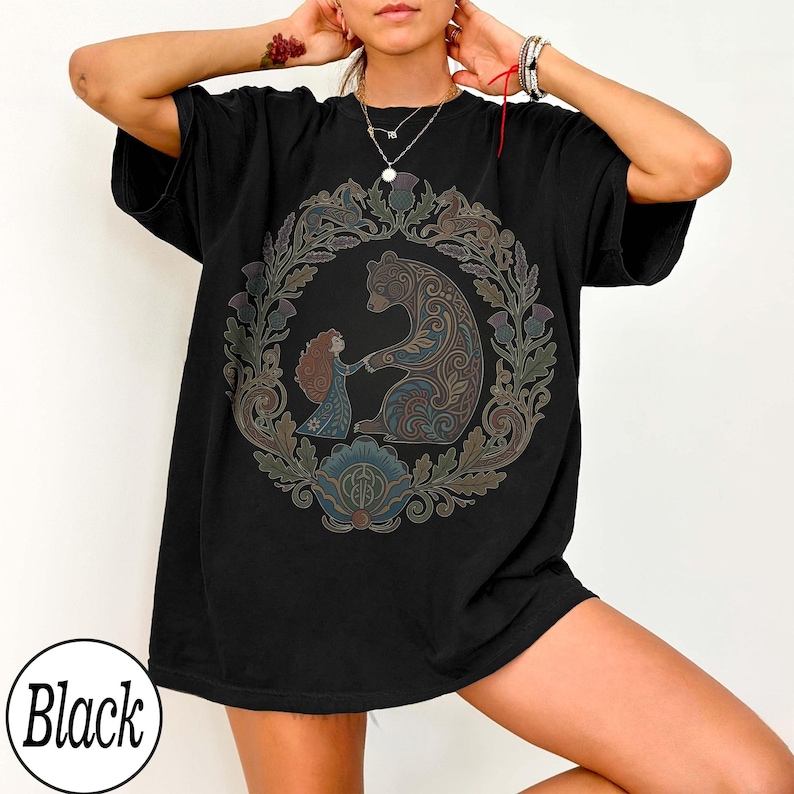 以下が含まれることがあります： 黒いTシャツ。円形のデザインで、クマと女の子が手をつないでおり、花のモチーフとアザミで囲まれています。デザインは茶色、青、緑の色合いを使用しています。「Black」の文字が下部の白い円の中にあります。