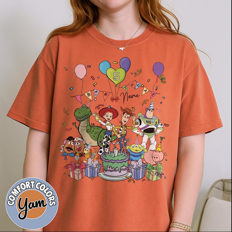 May include: Orange t-shirt featuring a colorful graphic of Toy Story characters celebrating a birthday. The design includes a birthday cake, balloons, and the number 5. The shirt has the Comfort Colors and Yam logos.