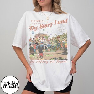 May include: White t-shirt featuring a colourful graphic of Toy Story Land in Florida. The design includes characters like Woody and Mickey Mouse, with the text "Toy Story Land" and "To Infinity and Beyond". The shirt has short sleeves.