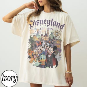 Puede incluir: Una camiseta de manga corta color marfil con un estampado gr&aacute;fico de personajes de Disney con disfraces de Halloween frente a un castillo. El texto dice Disneyland EST. 1955. Los personajes incluyen a Mickey, Minnie, Donald y Pluto.