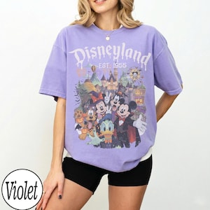 Puede incluir: Una camiseta violeta de manga corta con un estampado gr&aacute;fico de personajes de Disney con disfraces de Halloween frente a un castillo. El texto "Disneyland EST. 1955" est&aacute; impreso en blanco sobre los personajes. Tiene cuello redondo.