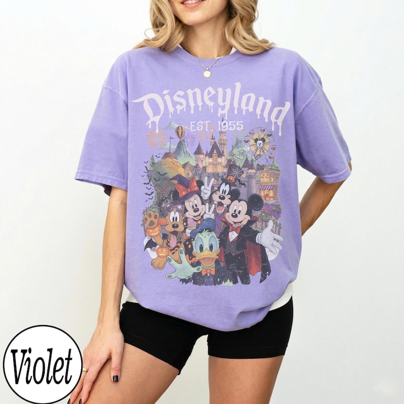 Puede incluir: Una camiseta violeta de manga corta con un estampado gr&aacute;fico de personajes de Disney con disfraces de Halloween frente a un castillo. El texto "Disneyland EST. 1955" est&aacute; impreso en blanco sobre los personajes. Tiene cuello redondo.