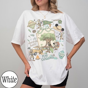 May include: White t-shirt with a vibrant graphic of cartoon characters, a hot air balloon, and the text "Adventure Awaits!". The design includes a tree, a car, and the words "Peak Challenge".
