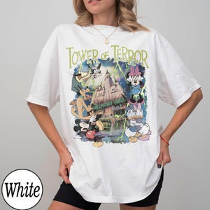 K&ouml;nnte beinhalten: Wei&szlig;es T-Shirt mit einer Grafik von Disney-Figuren Mickey Mouse, Goofy, Minnie Mouse und Donald und Daisy Duck vor dem Hollywood Tower Hotel. Der Text "Tower of Terror" ist in Gr&uuml;n &uuml;ber der Grafik aufgedruckt.