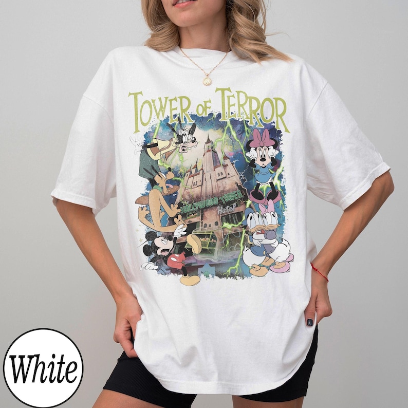 K&ouml;nnte beinhalten: Wei&szlig;es T-Shirt mit einer Grafik von Disney-Figuren Mickey Mouse, Goofy, Minnie Mouse und Donald und Daisy Duck vor dem Hollywood Tower Hotel. Der Text "Tower of Terror" ist in Gr&uuml;n &uuml;ber der Grafik aufgedruckt.