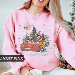 K&ouml;nnte beinhalten: Hellrosa Sweatshirt mit festlichem Aufdruck. Das Design zeigt Disney-Figuren in einem roten Auto, einen Weihnachtsbaum, Schneeflocken und einen Vollmond. Der Text "Merry Christmas" ist ebenfalls enthalten.