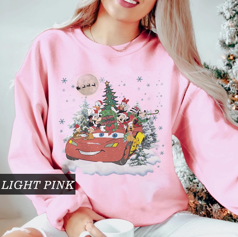 K&ouml;nnte beinhalten: Hellrosa Sweatshirt mit festlichem Aufdruck. Das Design zeigt Disney-Figuren in einem roten Auto, einen Weihnachtsbaum, Schneeflocken und einen Vollmond. Der Text "Merry Christmas" ist ebenfalls enthalten.