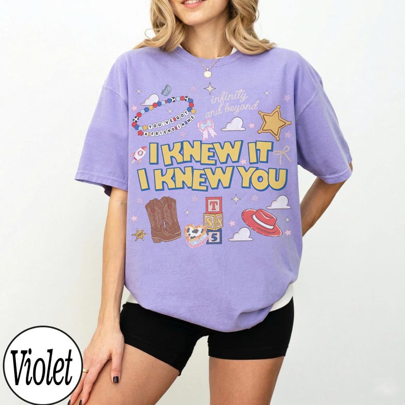 May include: A violet short-sleeve t-shirt featuring the yellow text "I KNEW IT I KNEW YOU" and various graphics including brown cowboy boots, a red hat, a gold star, alphabet blocks, and the phrases "infinity and beyond" and "you've got a friend in me."