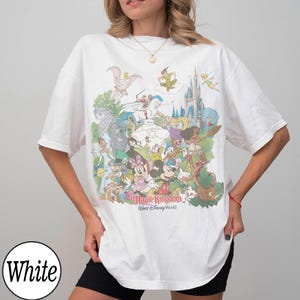 May include: White t-shirt featuring a colorful graphic of various Disney characters, including Mickey Mouse, Minnie Mouse, and Peter Pan. The text "Magic Kingdom, Walt Disney World" is printed below the characters. The shirt is a casual, oversized style.