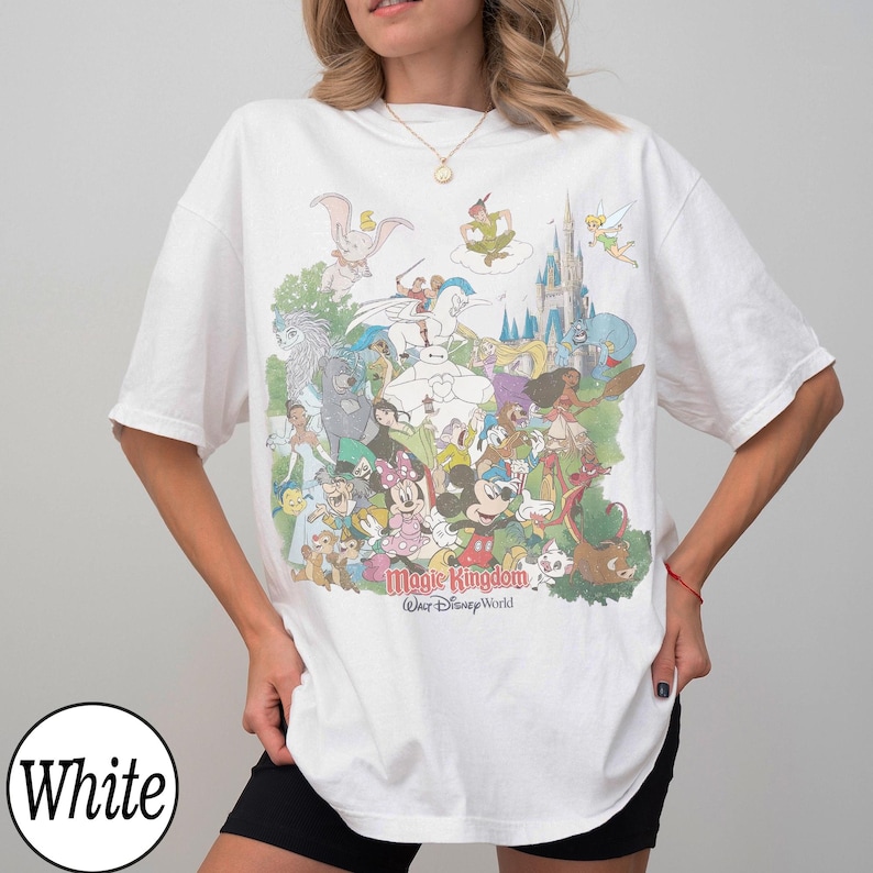 May include: White t-shirt featuring a colorful graphic of various Disney characters, including Mickey Mouse, Minnie Mouse, and Peter Pan. The text "Magic Kingdom, Walt Disney World" is printed below the characters. The shirt is a casual, oversized style.