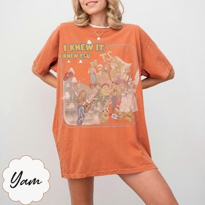 May include: An orange short-sleeve t-shirt featuring a graphic print of various animated characters and the text "I KNEW IT, I KNEW YOU" in yellow letters. The design includes a group of figures in a stylized scene with clouds and a star shape.