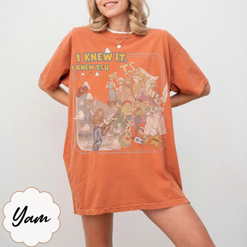 May include: An orange short-sleeve t-shirt featuring a graphic print of various animated characters and the text "I KNEW IT, I KNEW YOU" in yellow letters. The design includes a group of figures in a stylized scene with clouds and a star shape.