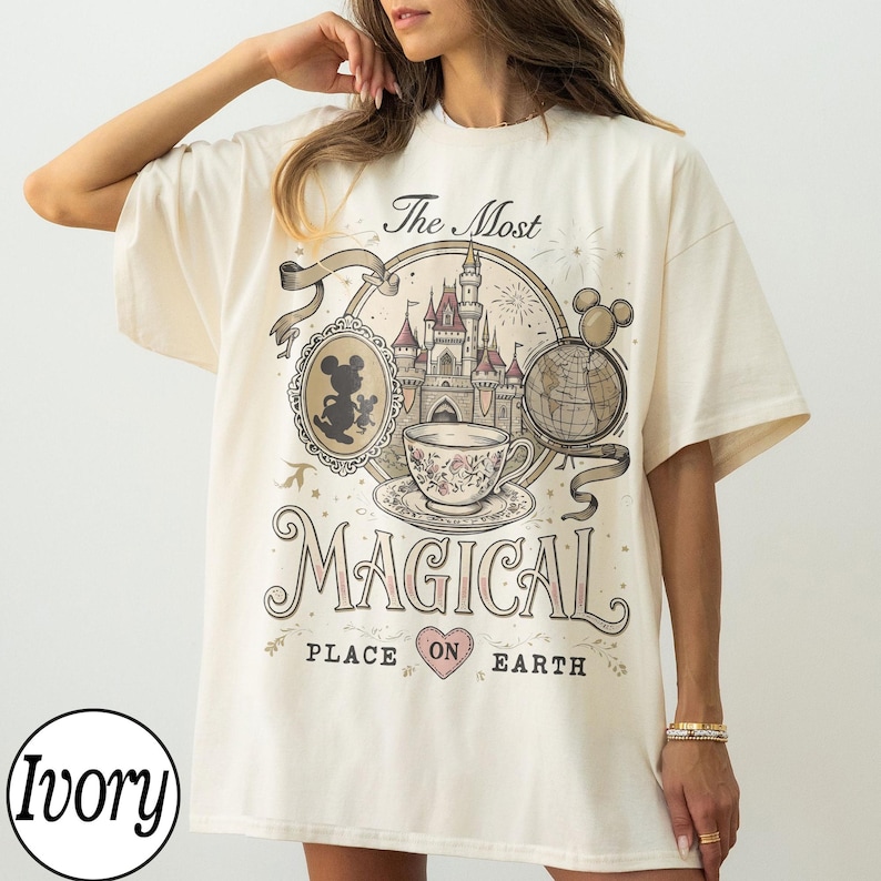 May include: Ivory-colored t-shirt with a graphic design featuring a castle, teacup, and Mickey Mouse silhouette. The text reads "The Most Magical Place on Earth." The shirt has short sleeves and a relaxed fit.