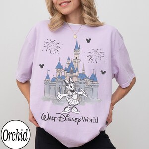 May include: A lilac t-shirt with a graphic of Cinderella's castle, fireworks, and Daisy Duck. The text "Walt Disney World" is printed below the graphic. The shirt also has the word "Orchid" printed on the bottom left.