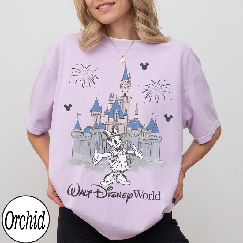 May include: A lilac t-shirt with a graphic of Cinderella's castle, fireworks, and Daisy Duck. The text "Walt Disney World" is printed below the graphic. The shirt also has the word "Orchid" printed on the bottom left.