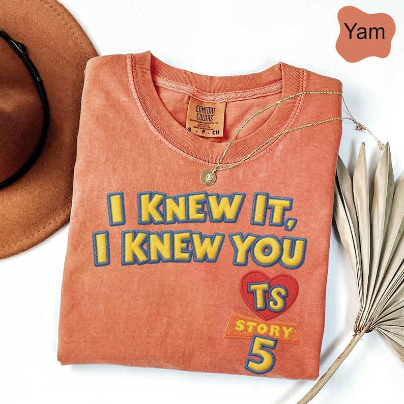 Puede incluir: Una camiseta de cuello redondo color &ntilde;ame con el texto bordado en amarillo y azul I KNEW IT, I KNEW YOU. Debajo del texto hay un coraz&oacute;n rojo con las letras TS, con la palabra STORY y el n&uacute;mero 5 impresos en amarillo debajo del coraz&oacute;n.