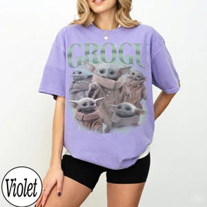 Puede incluir: Una camiseta violeta con un dise&ntilde;o de Grogu de The Mandalorian. El dise&ntilde;o incluye m&uacute;ltiples im&aacute;genes de Grogu, con la palabra "GROGU" en una fuente verde estilizada en la parte superior.