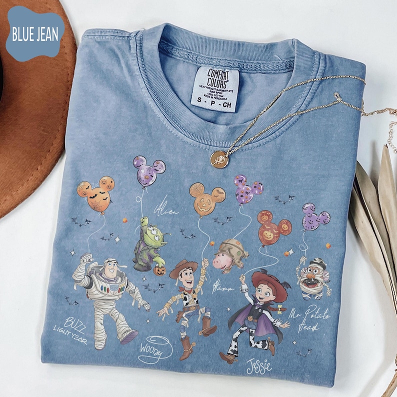 Pode incluir: Uma camiseta azul jeans Comfort Colors com personagens de Toy Story da Disney e bal&otilde;es do Mickey Mouse. A camisa inclui Buzz Lightyear, Woody, Jessie e Sr. Cabe&ccedil;a de Batata. Um colar de ouro est&aacute; sobre a camisa.