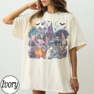 May include: An ivory t-shirt featuring a graphic print of two blue and pink alien characters dressed in Halloween costumes. One character is inside a pumpkin, and the other is wrapped in bandages like a mummy. The background shows a castle, bats, ghosts, and a crescent moon.