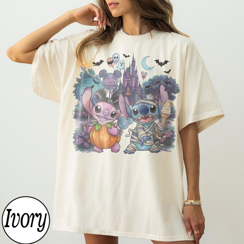 May include: An ivory t-shirt featuring a graphic print of two blue and pink alien characters dressed in Halloween costumes. One character is inside a pumpkin, and the other is wrapped in bandages like a mummy. The background shows a castle, bats, ghosts, and a crescent moon.