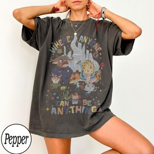 May include: Dark grey t-shirt with a colourful graphic of animated animal characters and the text "WHERE ANYONE CAN BE ANYTHING". The design includes a cityscape and various cartoon figures.