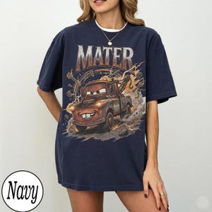 May include: Navy blue t-shirt featuring a graphic of Mater from the movie Cars. The design includes the word "MATER" in a metallic font above an illustration of the character. The shirt is a relaxed fit.