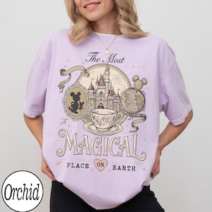 May include: A light purple t-shirt featuring a whimsical design with a castle, teacup, and globe. The text reads "The Most Magical Place on Earth." A silhouette of Mickey Mouse is in a cameo. The brand name "Orchid" is at the bottom.