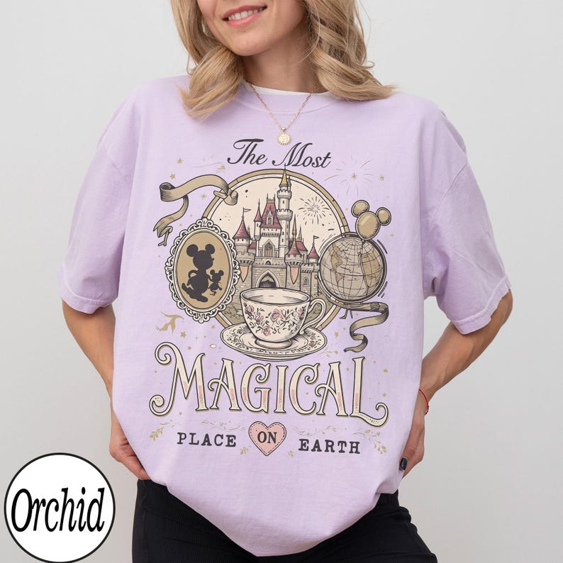 May include: A light purple t-shirt featuring a whimsical design with a castle, teacup, and globe. The text reads "The Most Magical Place on Earth." A silhouette of Mickey Mouse is in a cameo. The brand name "Orchid" is at the bottom.