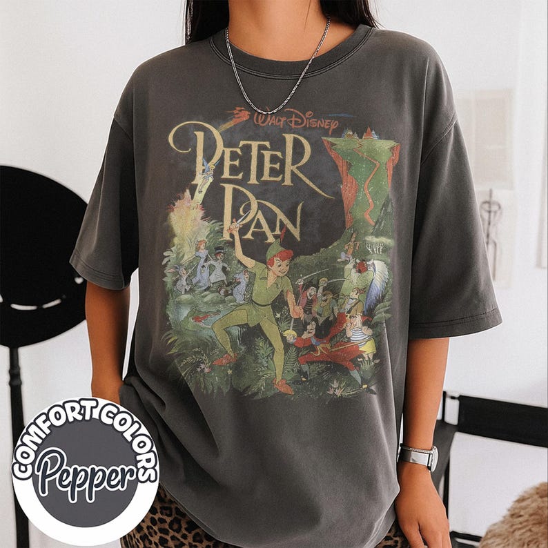 May include: Dark gray Comfort Colors t-shirt featuring a vintage-style Peter Pan graphic. The design includes the text "Walt Disney Peter Pan" with illustrations of Peter Pan, Captain Hook, and other characters from the movie.