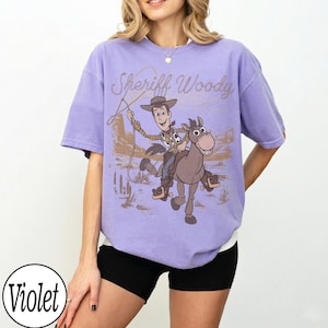 May include: A violet-colored t-shirt featuring a graphic of Sheriff Woody riding a horse, with the text "Sheriff Woody" above the image. The design is in shades of brown and tan, set against a desert backdrop.