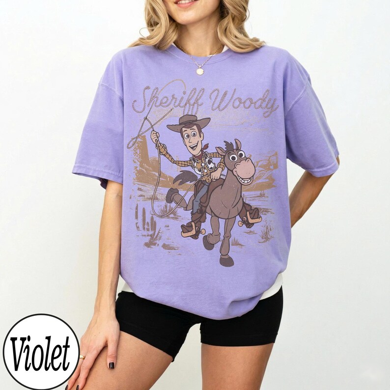 May include: A violet-colored t-shirt featuring a graphic of Sheriff Woody riding a horse, with the text "Sheriff Woody" above the image. The design is in shades of brown and tan, set against a desert backdrop.