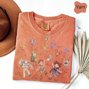Pode incluir: Uma camiseta Comfort Colors de gola redonda cor inhame com um gr&aacute;fico de personagens de Toy Story segurando bal&otilde;es em forma de Mickey Mouse. A camisa tem um colar de ouro e uma pequena etiqueta que diz "Comfort Colors".