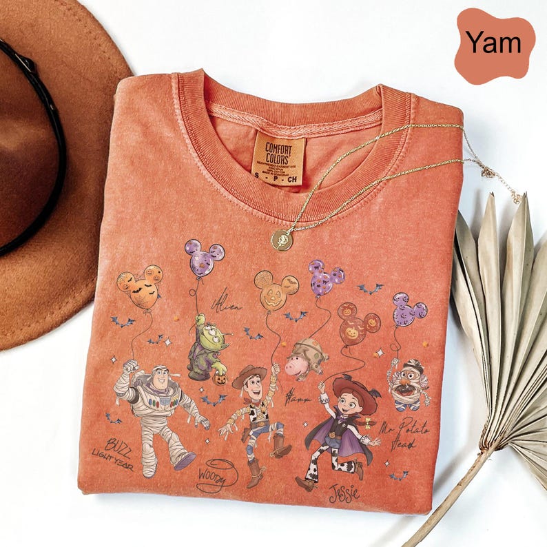 Pode incluir: Uma camiseta Comfort Colors de gola redonda cor inhame com um gr&aacute;fico de personagens de Toy Story segurando bal&otilde;es em forma de Mickey Mouse. A camisa tem um colar de ouro e uma pequena etiqueta que diz "Comfort Colors".