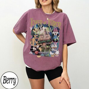 K&ouml;nnte beinhalten: Beerenfarbenes T-Shirt mit einer Grafik von Disney-Figuren und dem Text "Tower of Terror". Das Design zeigt Micky Maus, Minnie Maus, Donald Duck, Daisy Duck und Goofy vor dem Hollywood Tower Hotel.