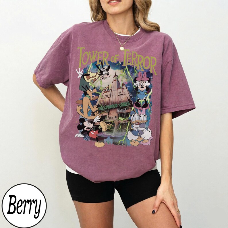 K&ouml;nnte beinhalten: Beerenfarbenes T-Shirt mit einer Grafik von Disney-Figuren und dem Text "Tower of Terror". Das Design zeigt Micky Maus, Minnie Maus, Donald Duck, Daisy Duck und Goofy vor dem Hollywood Tower Hotel.