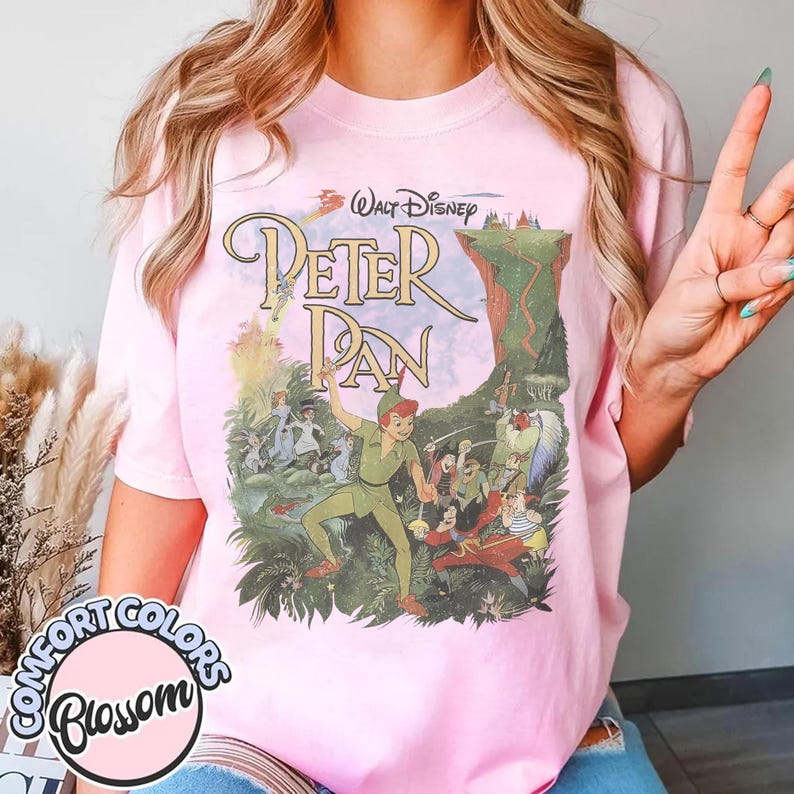 May include: Pink t-shirt featuring a vintage-style graphic of Peter Pan. The design includes the text "Walt Disney Peter Pan" and illustrations of characters from the movie. The shirt has a Comfort Colors Blossom label.