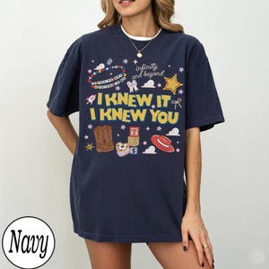 May include: A navy blue short-sleeve t-shirt featuring a graphic print with the text "I KNEW IT I KNEW YOU" and "infinity and beyond". The design includes illustrations of brown cowboy boots, a red hat, alphabet blocks, a star, and a beaded bracelet.