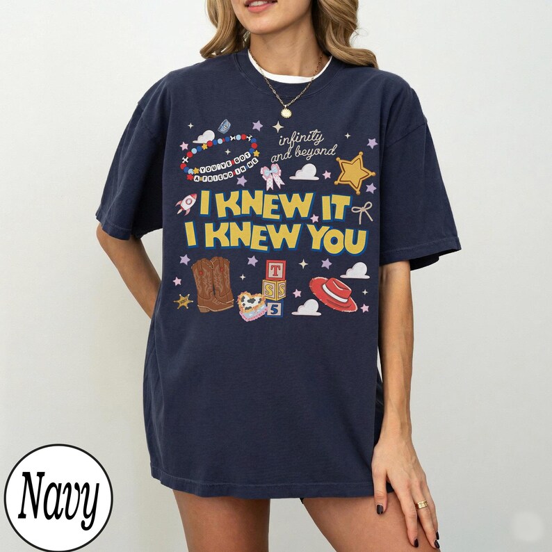 May include: A navy blue short-sleeve t-shirt featuring a graphic print with the text "I KNEW IT I KNEW YOU" and "infinity and beyond". The design includes illustrations of brown cowboy boots, a red hat, alphabet blocks, a star, and a beaded bracelet.