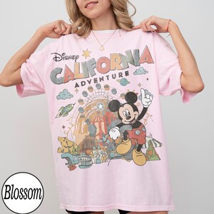 May include: A light pink t-shirt featuring the text "Disney CALIFORNIA ADVENTURE" with a graphic of Mickey Mouse and amusement park imagery. The shirt has short sleeves and a crew neck.