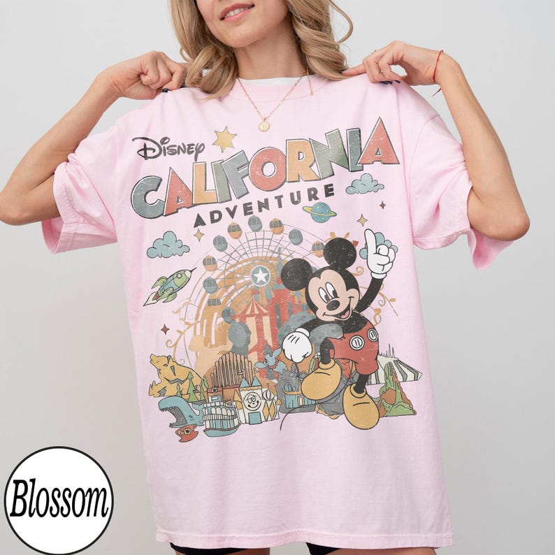 May include: A light pink t-shirt featuring the text "Disney CALIFORNIA ADVENTURE" with a graphic of Mickey Mouse and amusement park imagery. The shirt has short sleeves and a crew neck.
